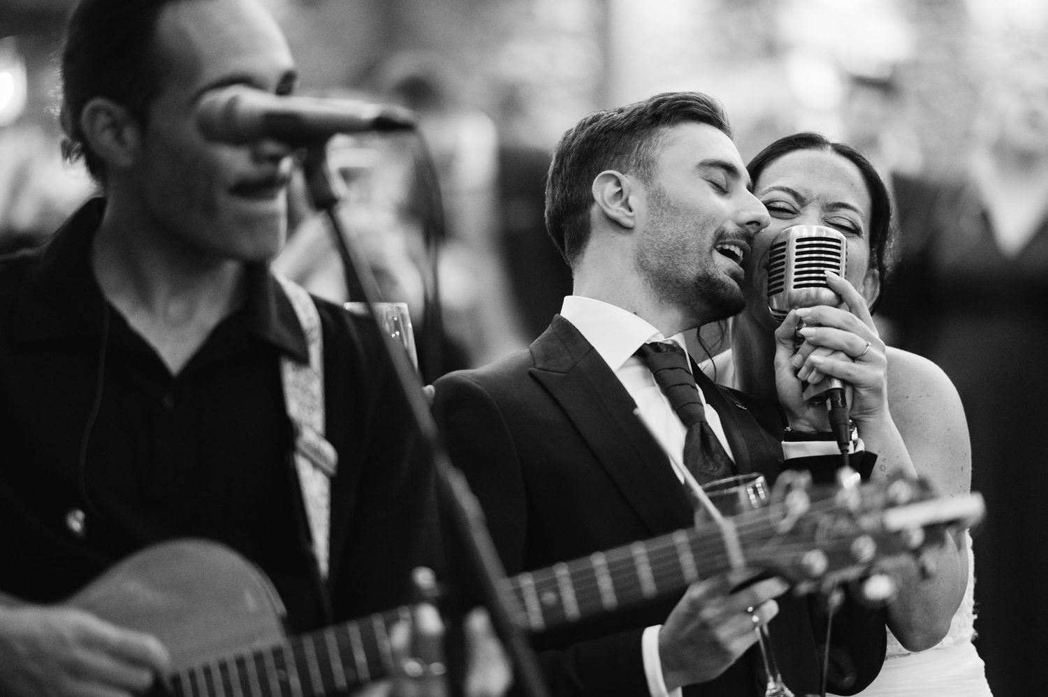 Musicians perform at a wedding reception with a guitar player and vocalist singing into a vintage microphone.