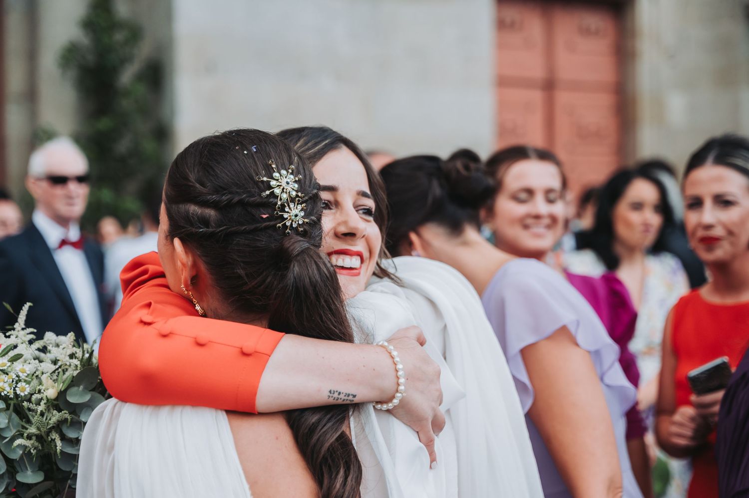Wedding party and guests celebrating with raised hands outside a venue with coral-colored doors.