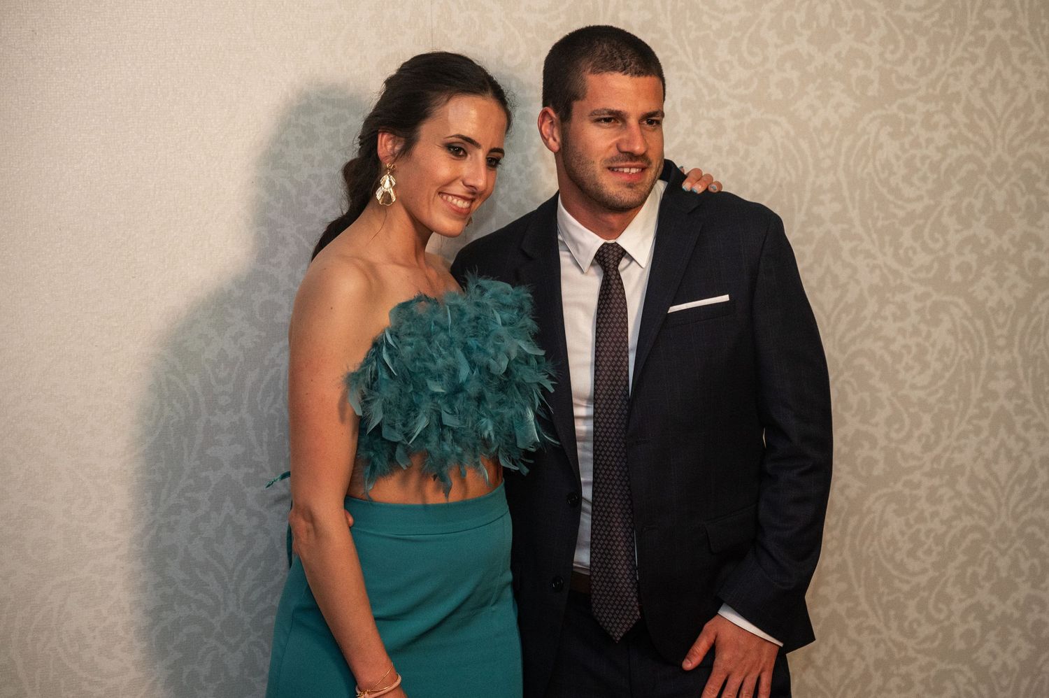 Formal event attendees in elegant attire pose together against a textured beige wall at a celebration.