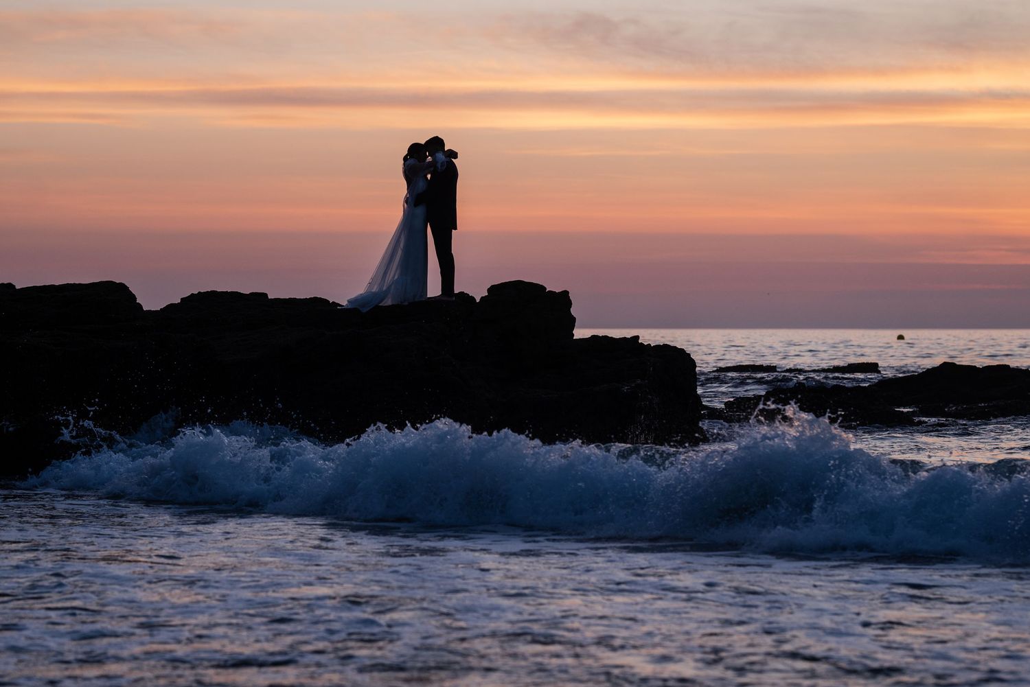 Silhouetted figures stand on rocky coastal outcrop at sunset as waves crash against the shore.