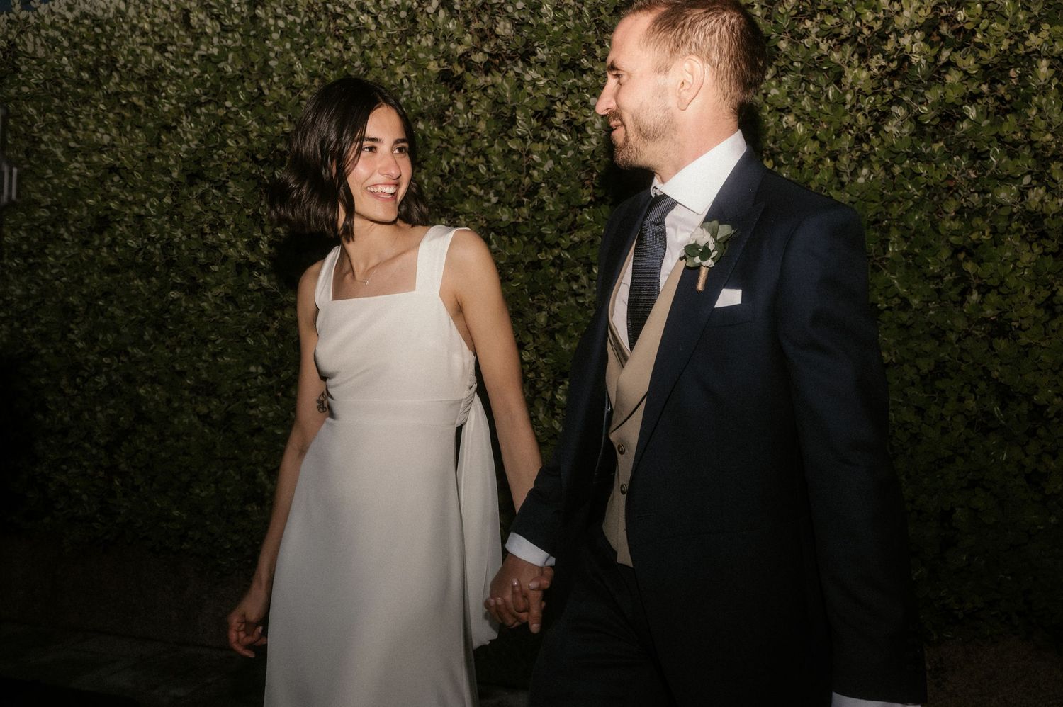 A couple in wedding attire walks together in front of a lush green hedge backdrop.