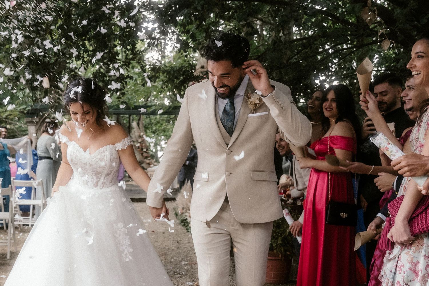 Outdoor wedding ceremony under trees with guests in formal attire and red dresses.