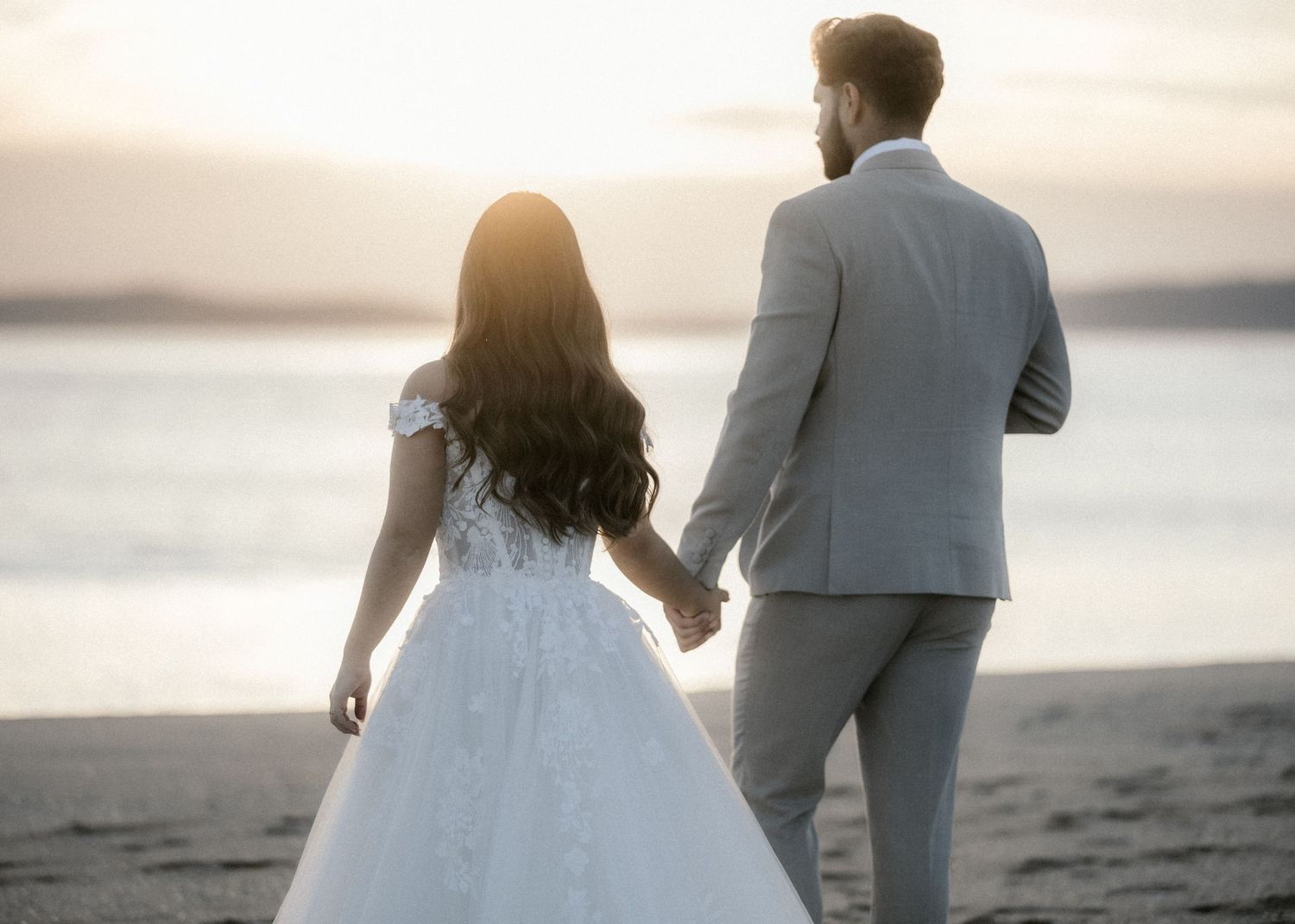 A couple in wedding attire holds hands while walking along a beach at sunset.