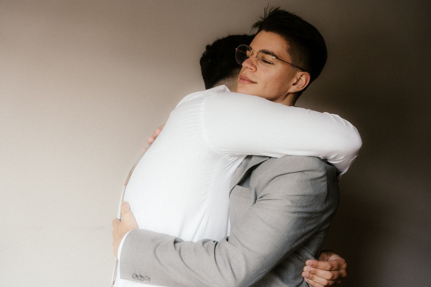 Two people embrace in an emotional hug, one wearing a light gray suit against a neutral background.