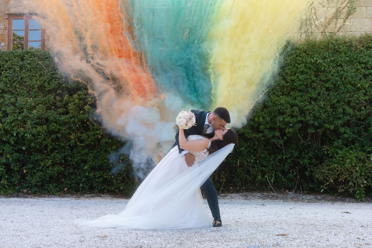A couple in wedding attire dips dramatically against a backdrop of colorful smoke bombs creating a rainbow effect.