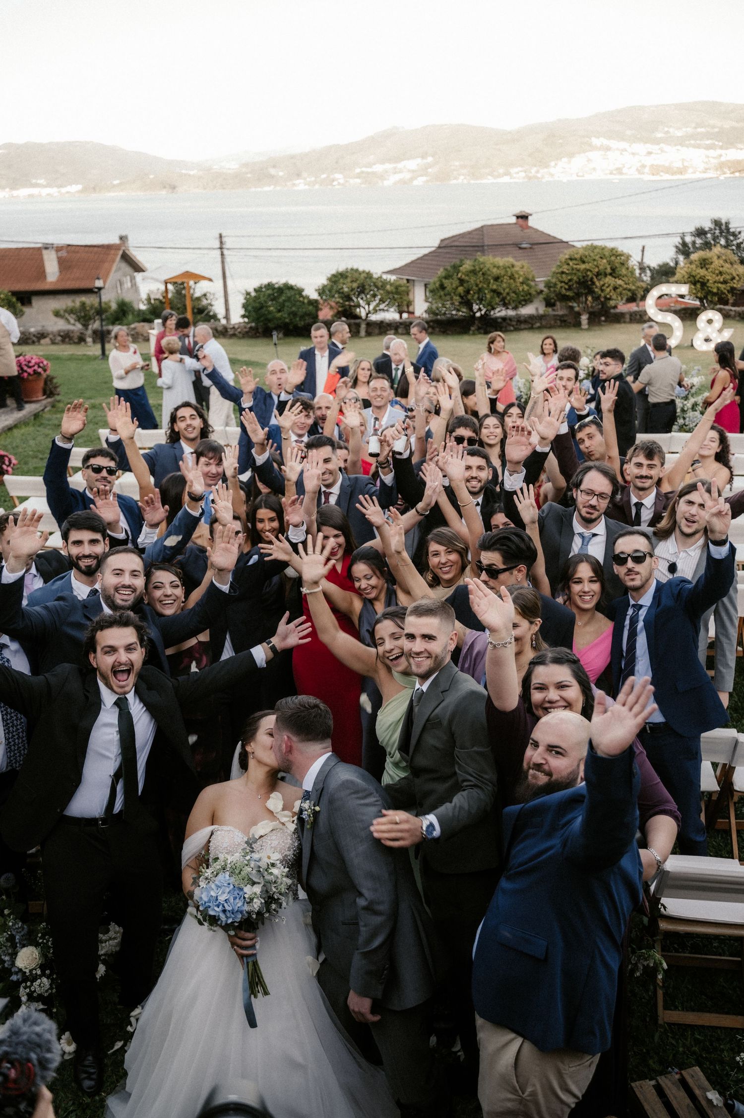 Wedding guests and newlyweds celebrate with raised hands and cheers at an outdoor waterfront venue.