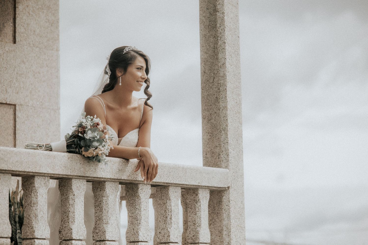 A bride in a white wedding dress poses elegantly against a stone pillar on a cloudy day.