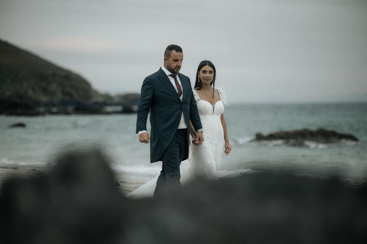A couple in wedding attire stands together on a moody rocky beach with overcast skies and ocean waves in the background.