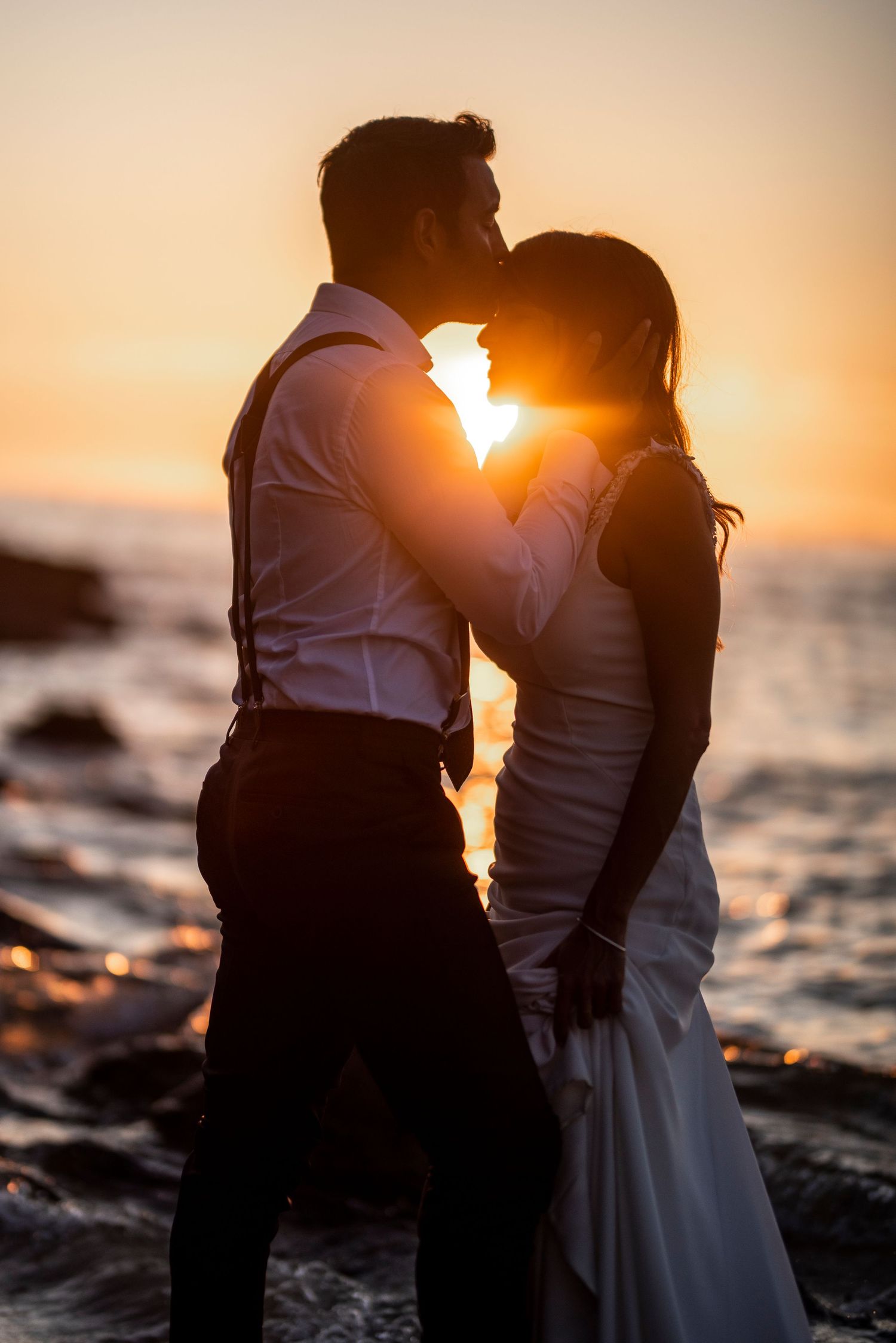 Silhouettes of a couple embrace against a dramatic ocean sunset with waves in the background.