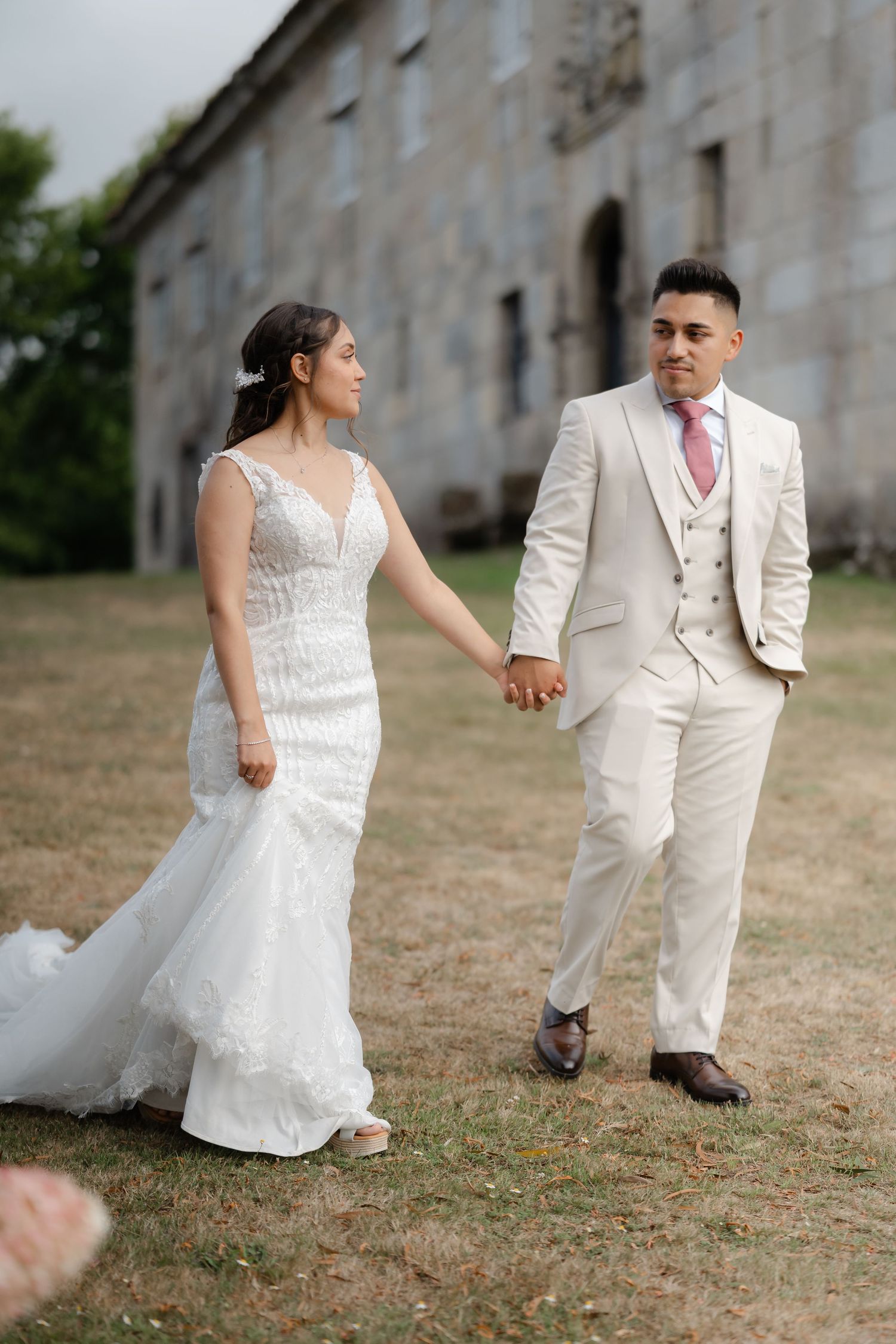 A couple in wedding attire holds hands while walking together on grass near a stone wall.