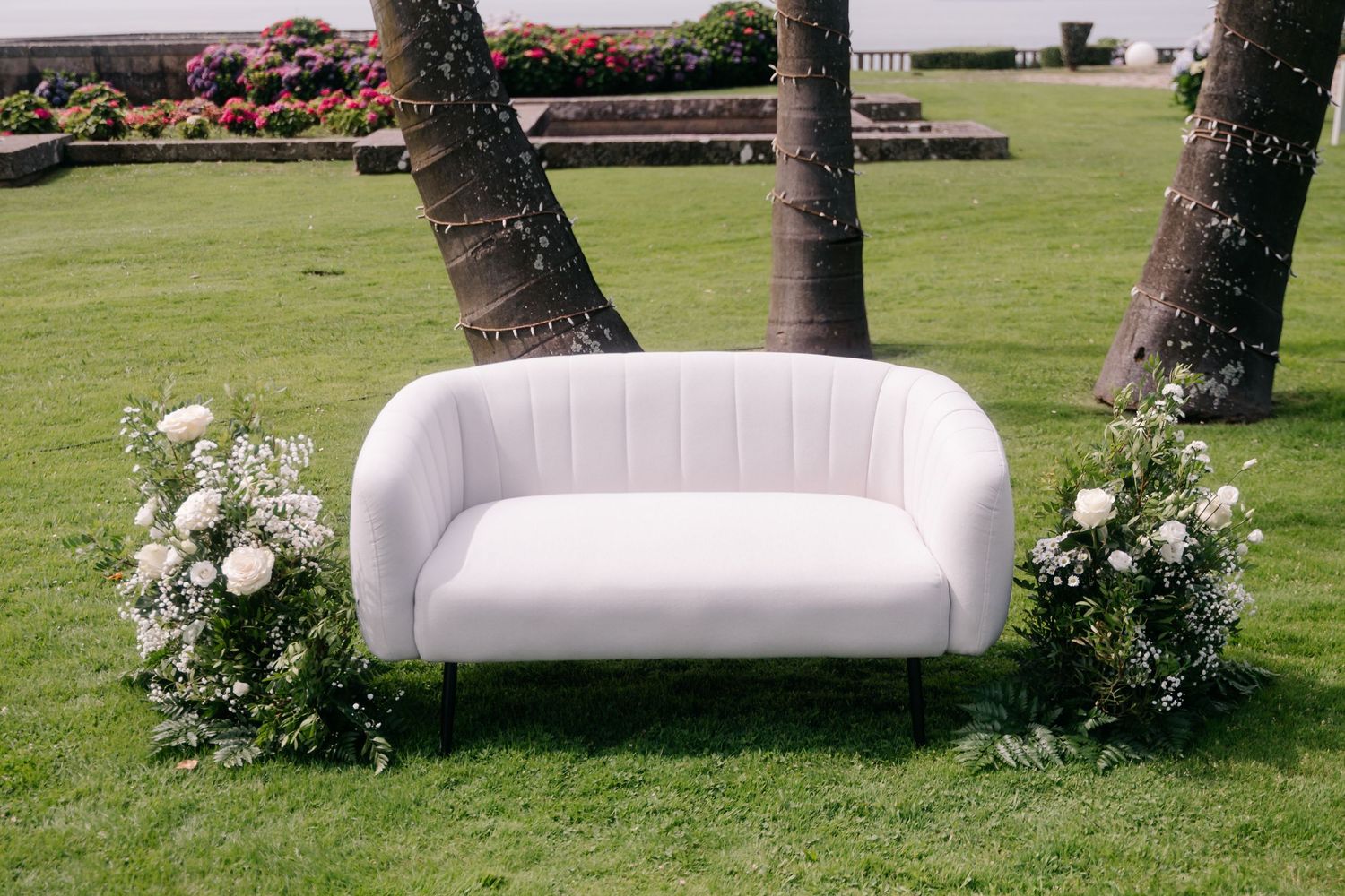 White velvet loveseat positioned on green lawn between two palm trees and white floral arrangements.