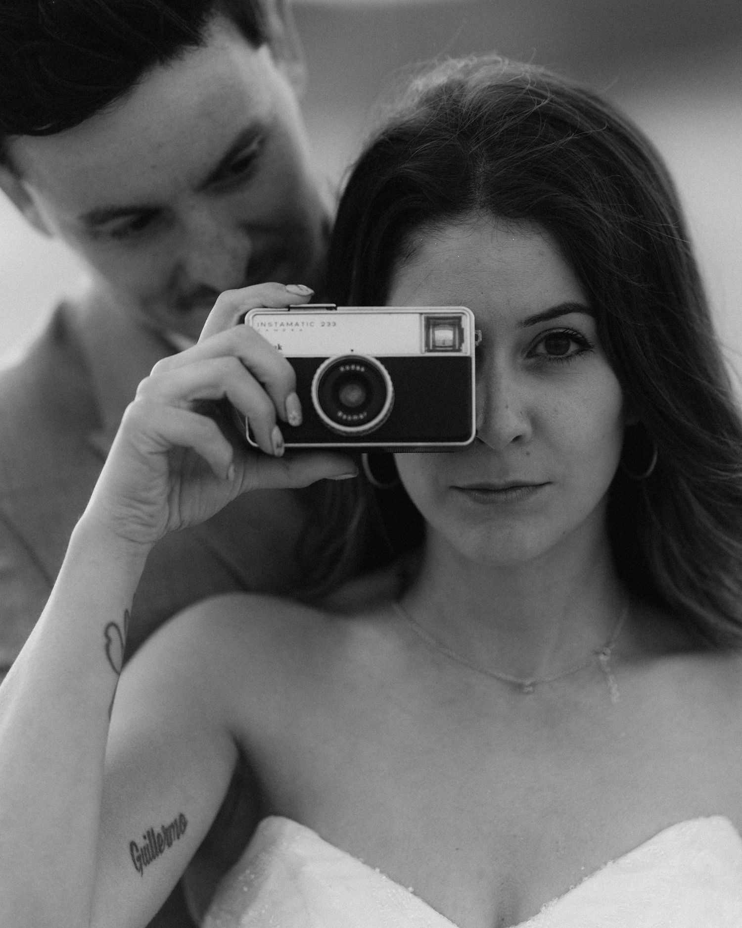 Black and white close-up of a bride holding a vintage camera up to her eye with groom behind her.