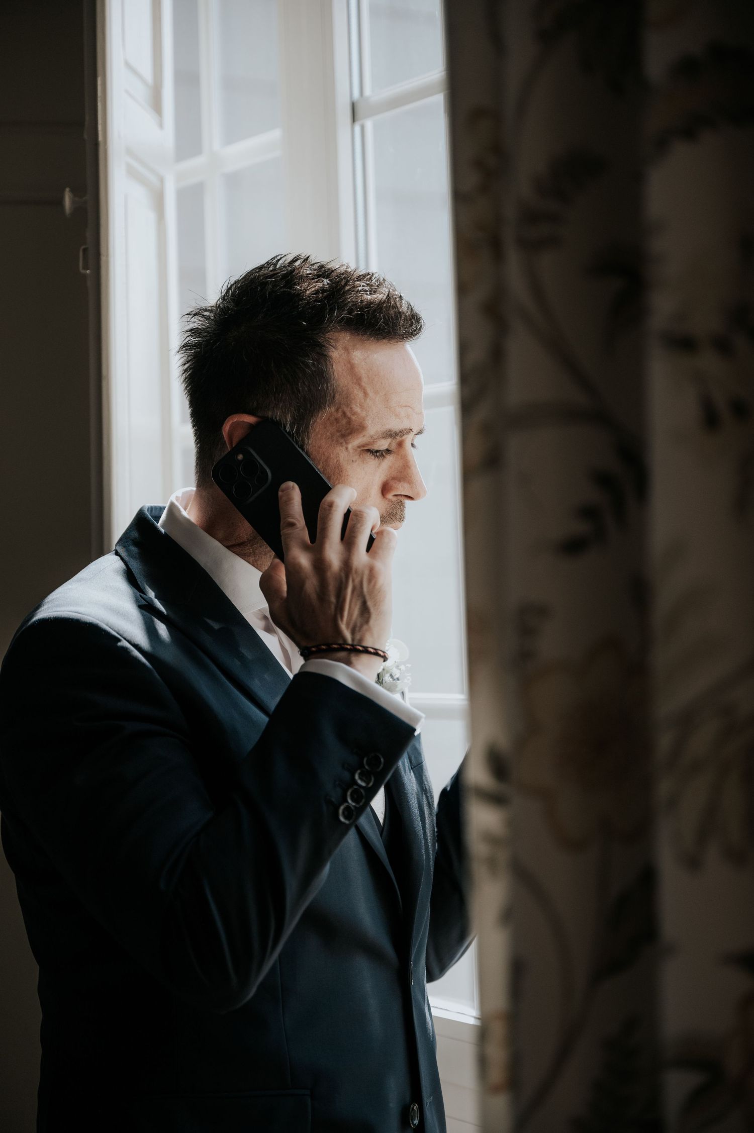 A person in a dark coat stands by a window speaking on a mobile phone in dramatic lighting.
