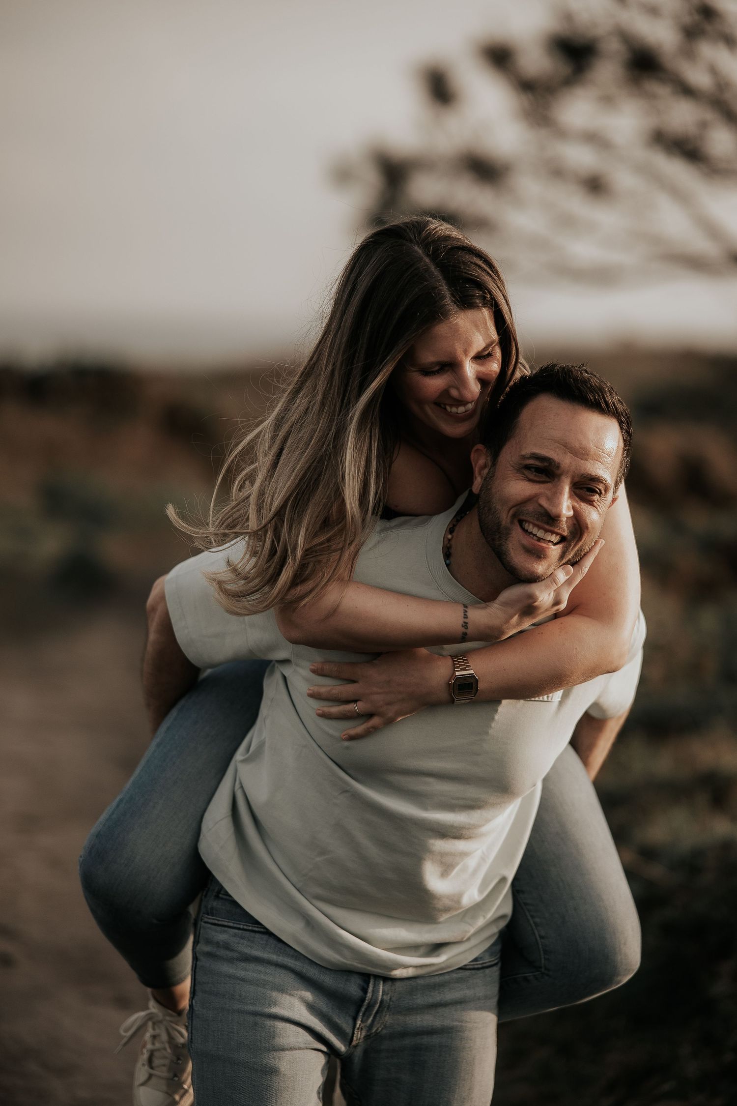 A playful couple laughs together during a piggyback ride outdoors in warm lighting.