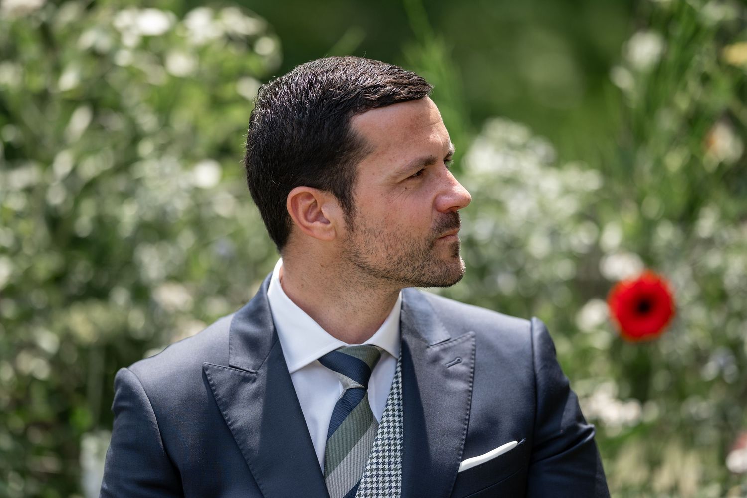 Groom in navy suit and grey tie stands against lush garden backdrop.