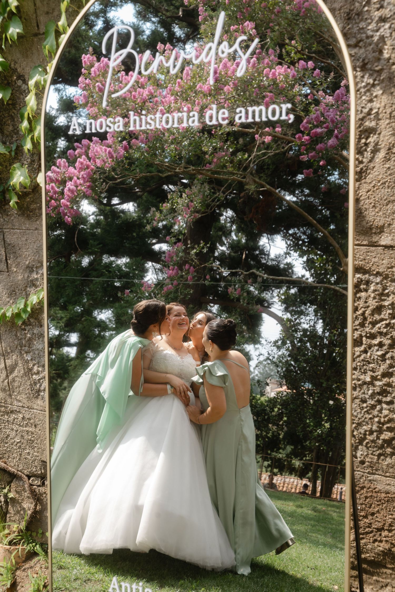 Bridesmaids in mint green dresses pose playfully with the bride in a white gown against a garden backdrop.