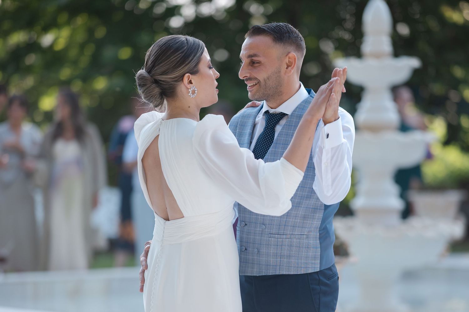 A couple in formal attire dances together outdoors in a romantic garden setting during a wedding celebration.