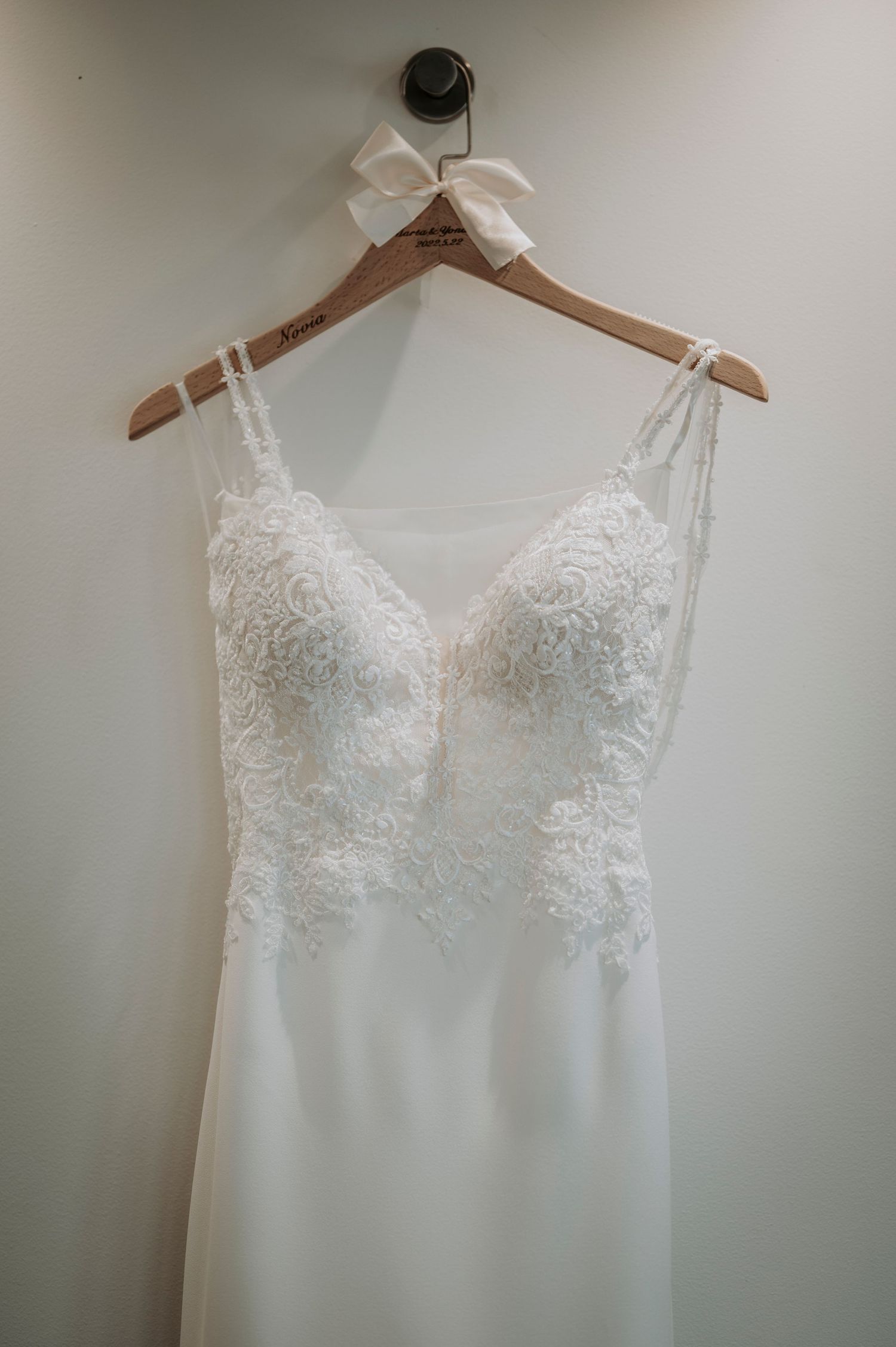White lace wedding dress with delicate floral details hangs elegantly on a wooden hanger against a light background.