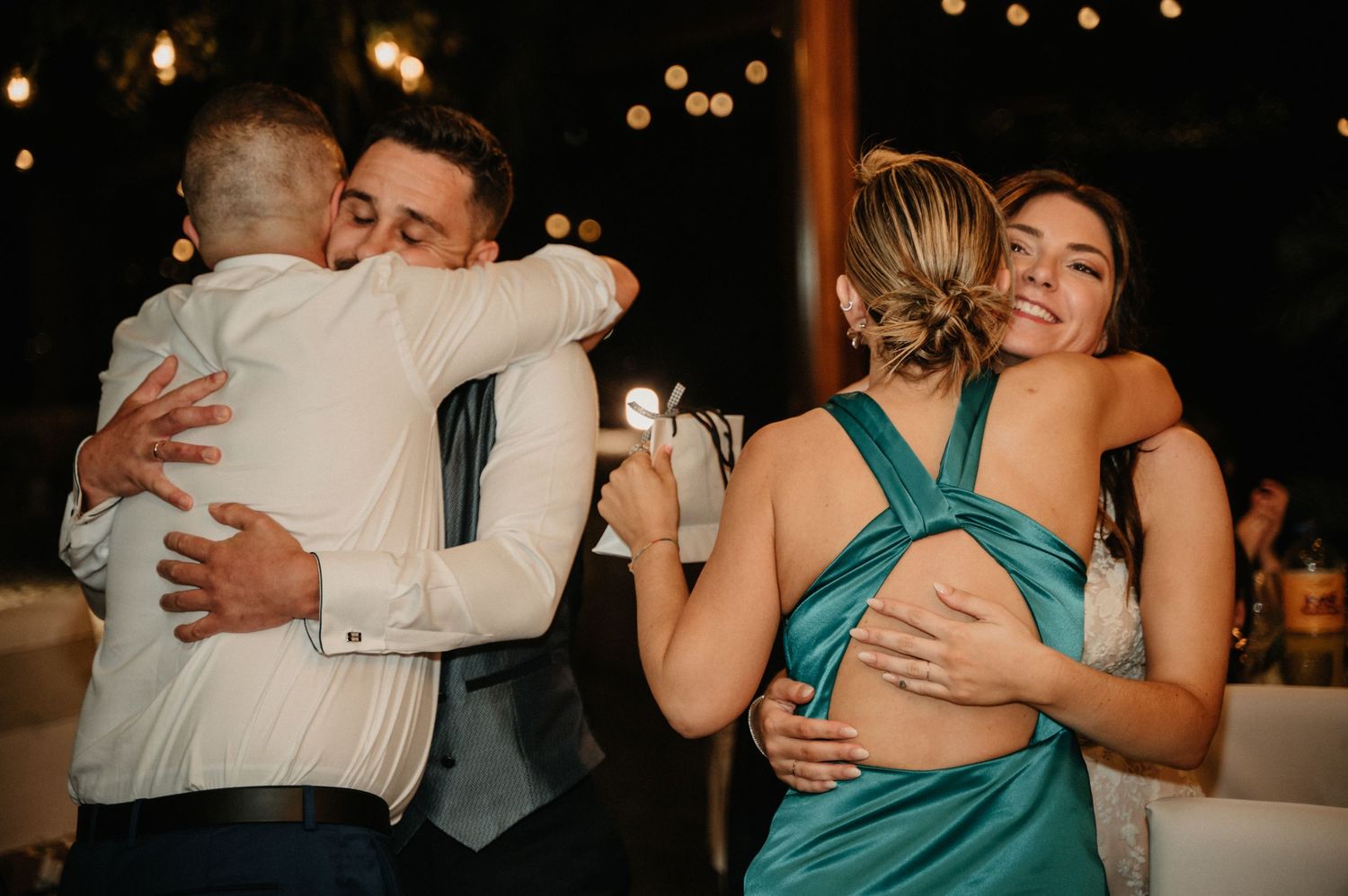 People embrace warmly at an evening celebration with glowing lights in the background.