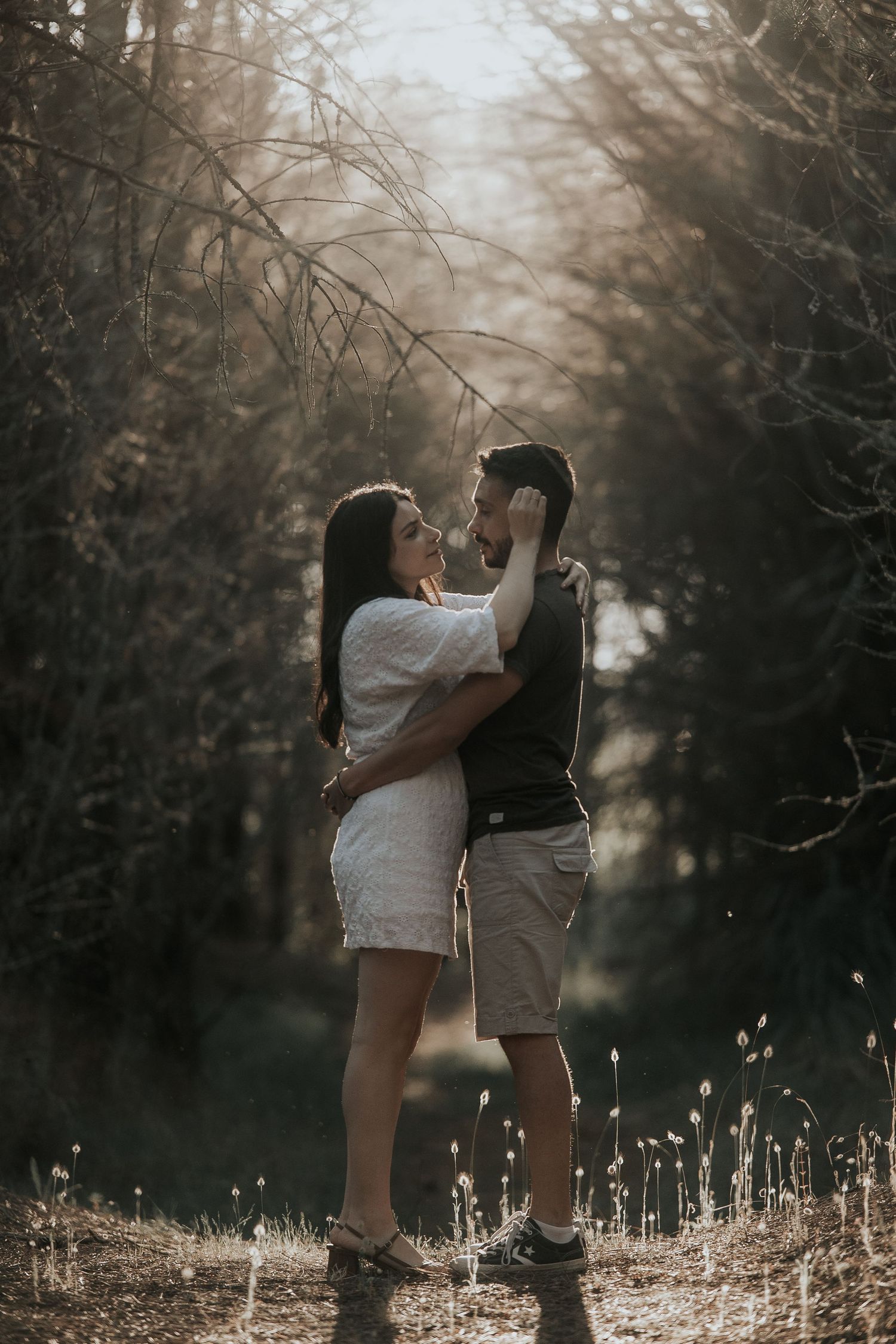 A couple embraces in a moody forest setting with sunlight filtering through bare trees and dried grass below.
