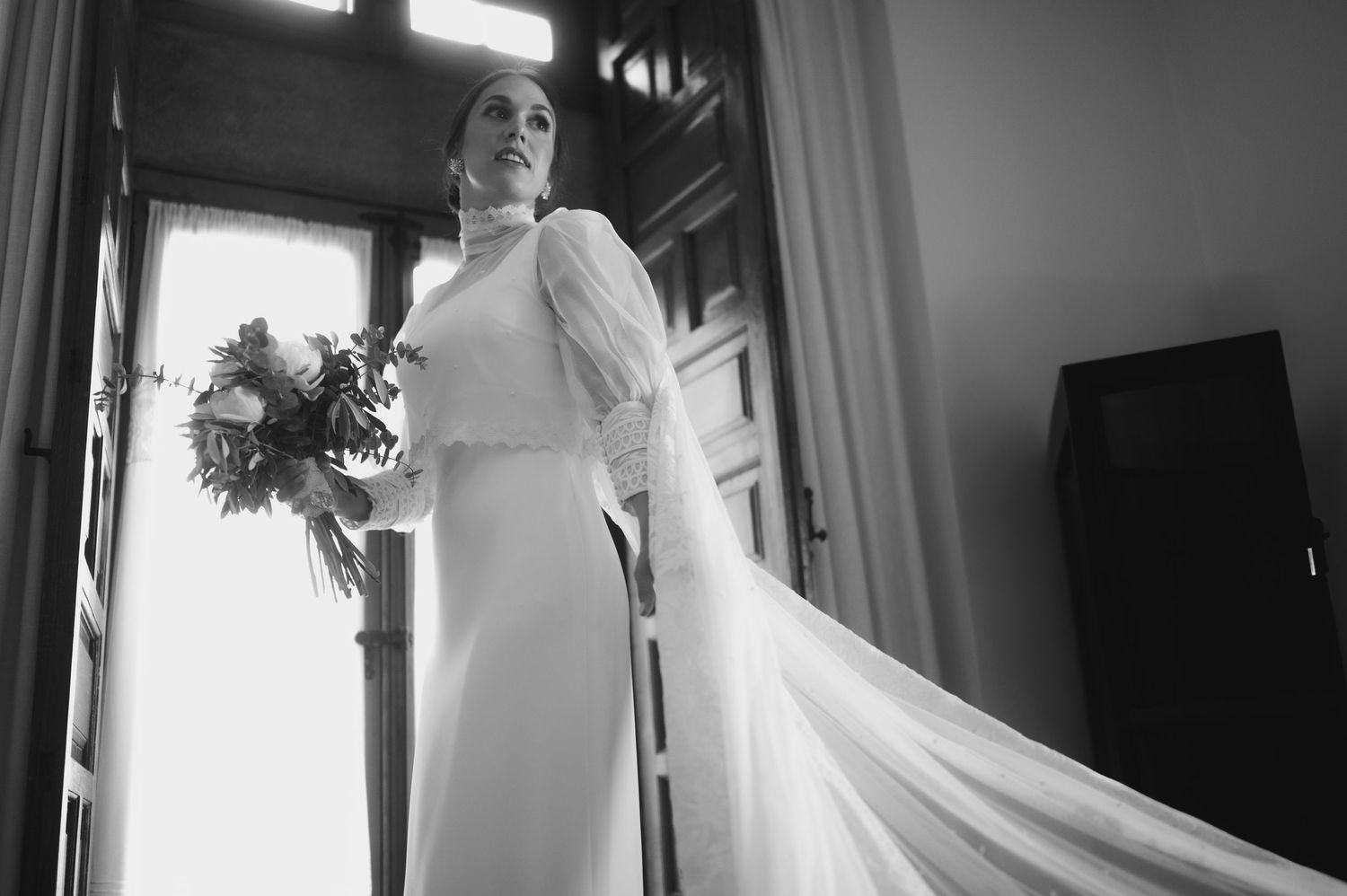 Black and white wedding photo showing a bride in a flowing white gown standing near a window in dramatic lighting.