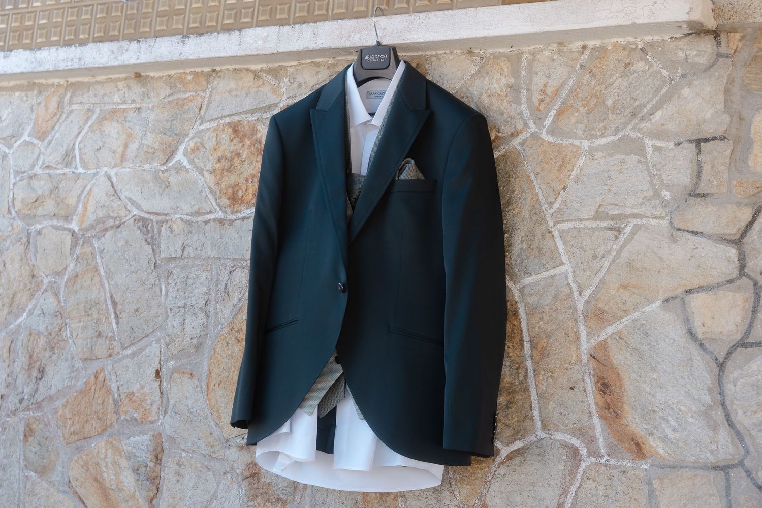 A dark green suit jacket with a pocket square hangs against a stone wall next to a white dress shirt.