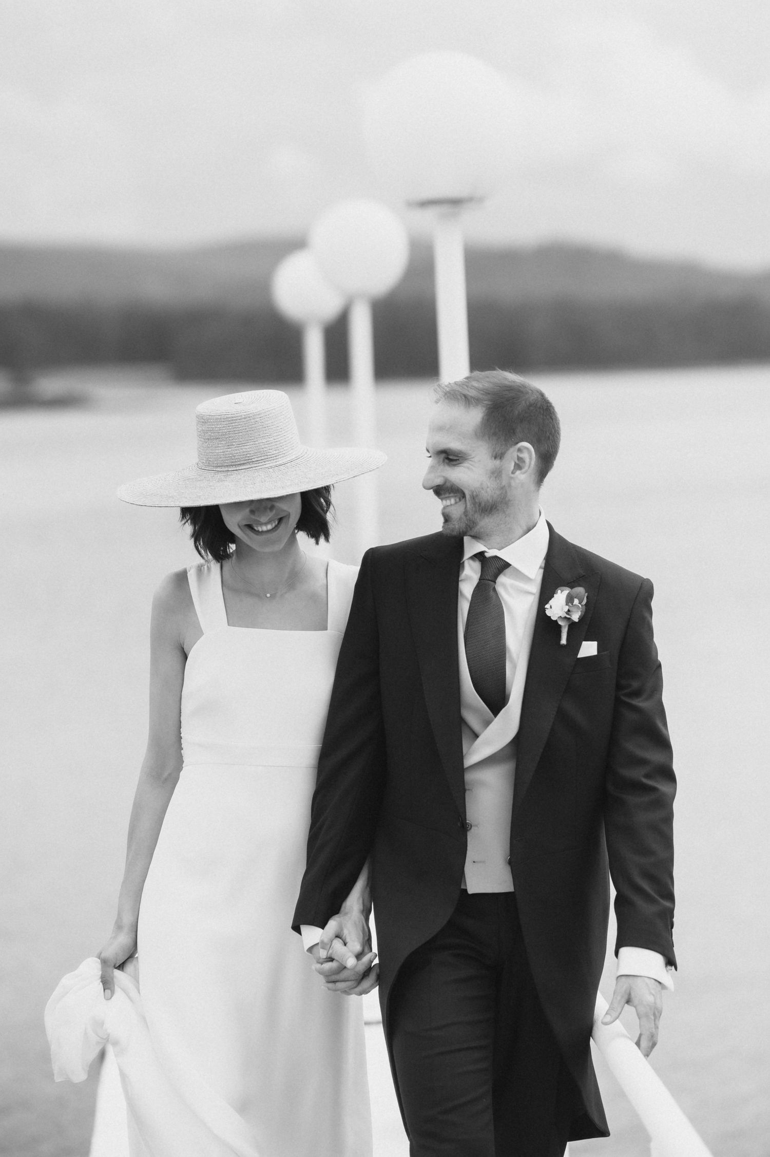 A romantic black and white wedding photo of a couple walking together by a lakeside wearing formal attire.