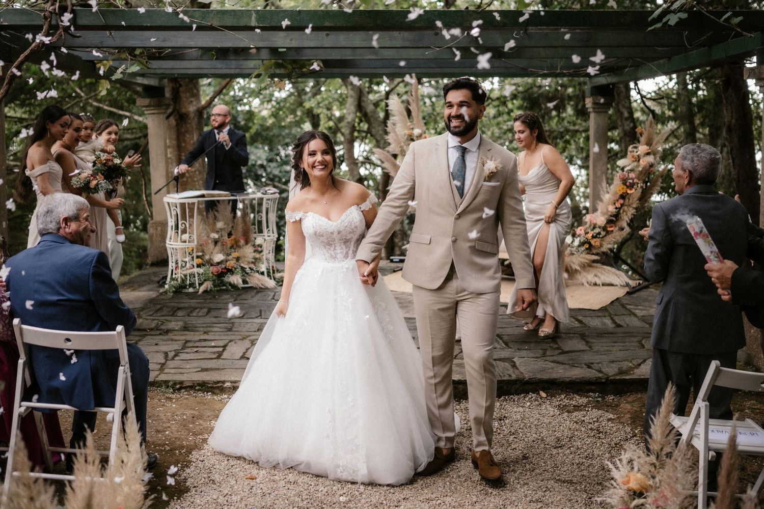 Outdoor wedding ceremony with guests seated on wooden benches surrounded by trees and natural lighting.