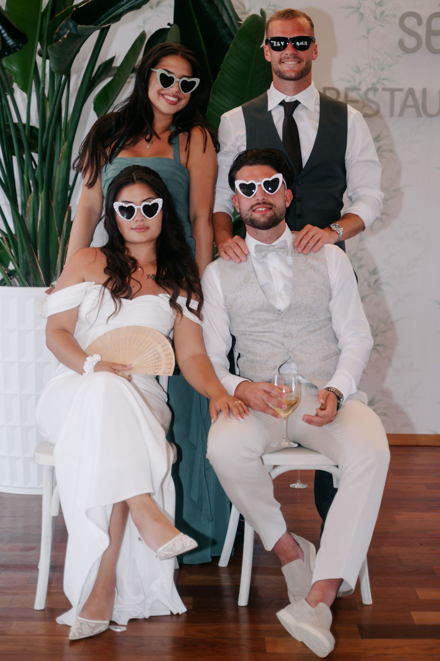 A stylish group wearing sunglasses and formal attire poses together in front of tropical plants and a light wall.