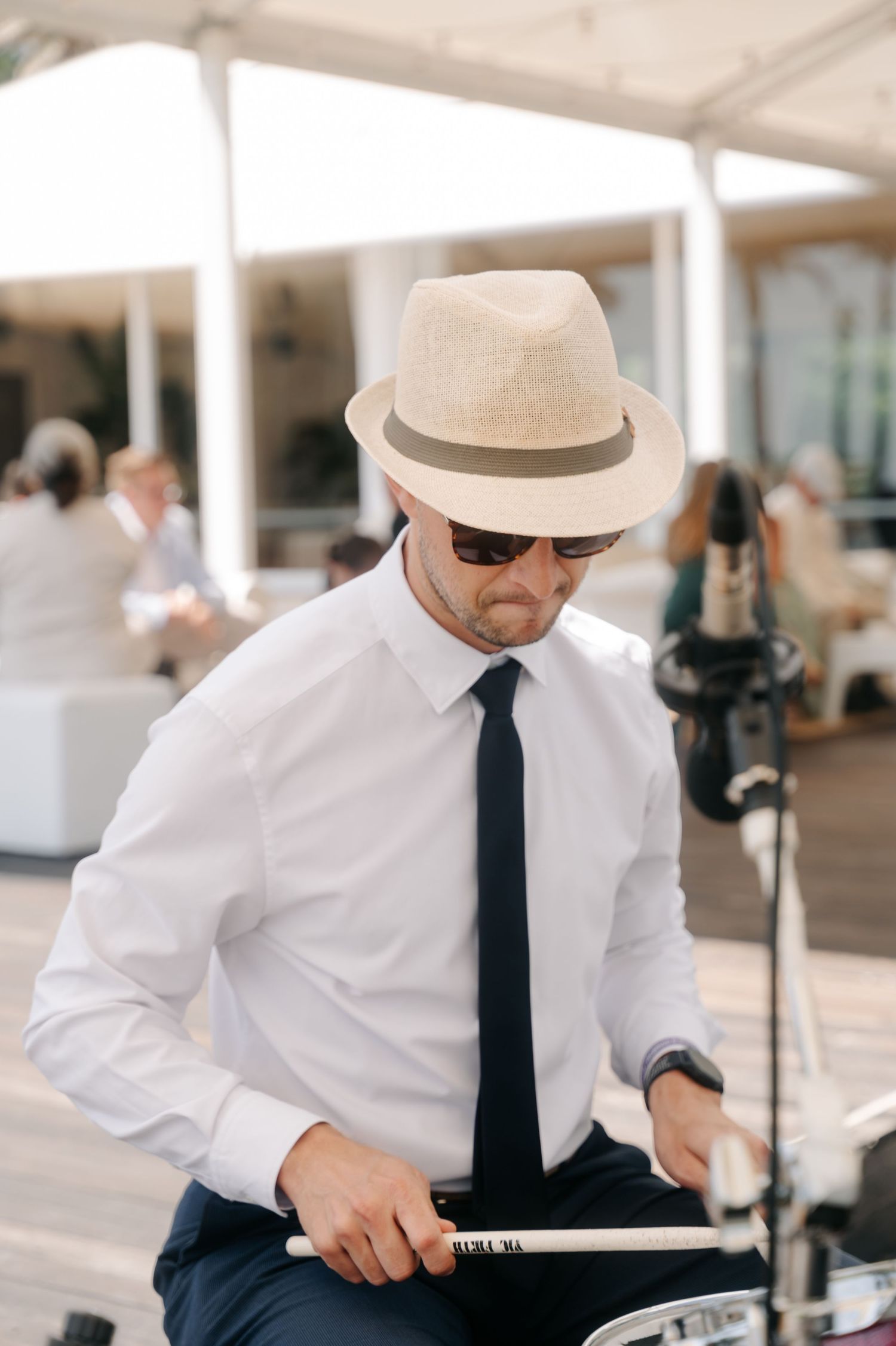 A person in a white shirt, black tie and fedora hat sits at a table outdoors.