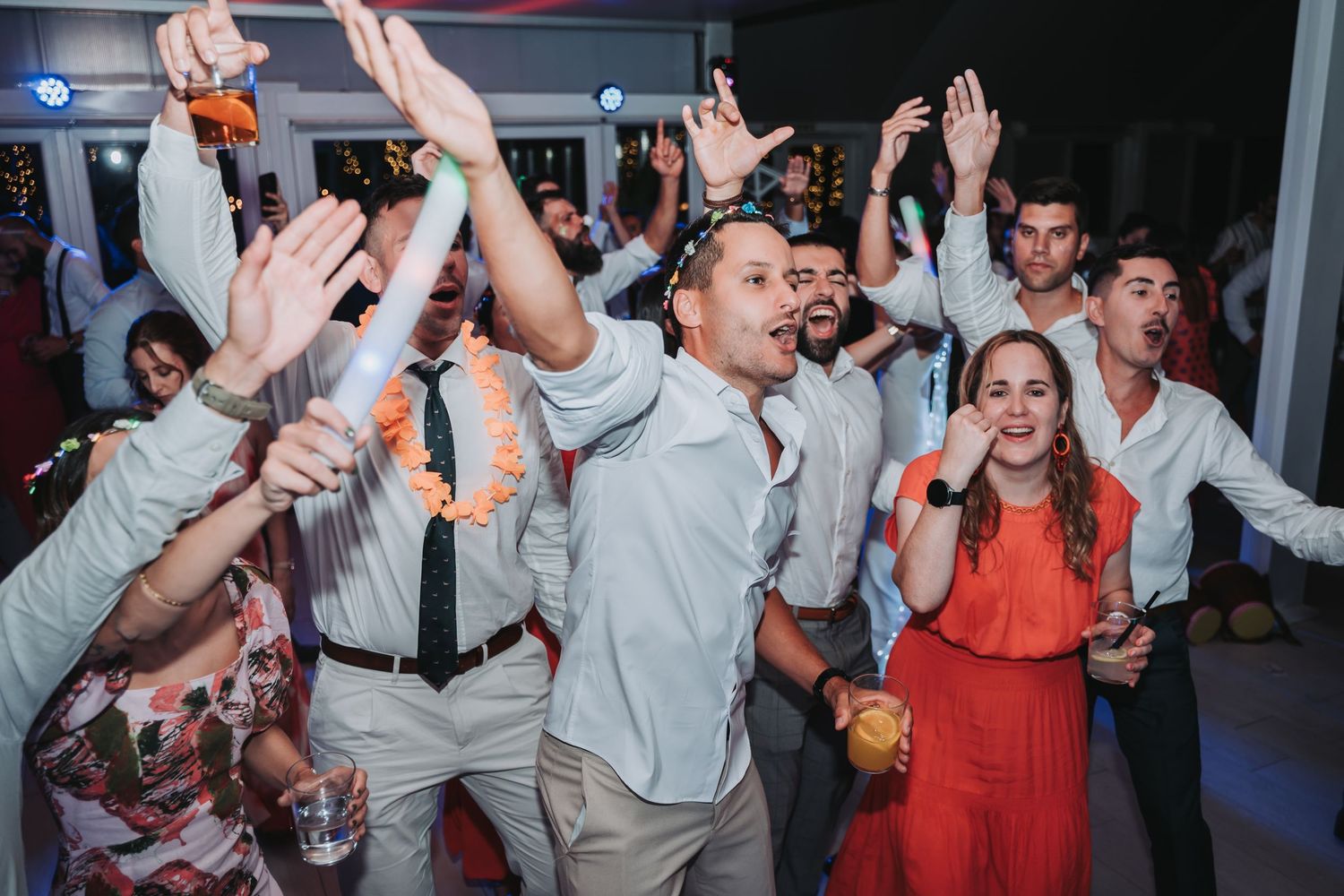 Energetic party crowd dancing and celebrating with raised arms at a festive indoor event with colorful lighting.
