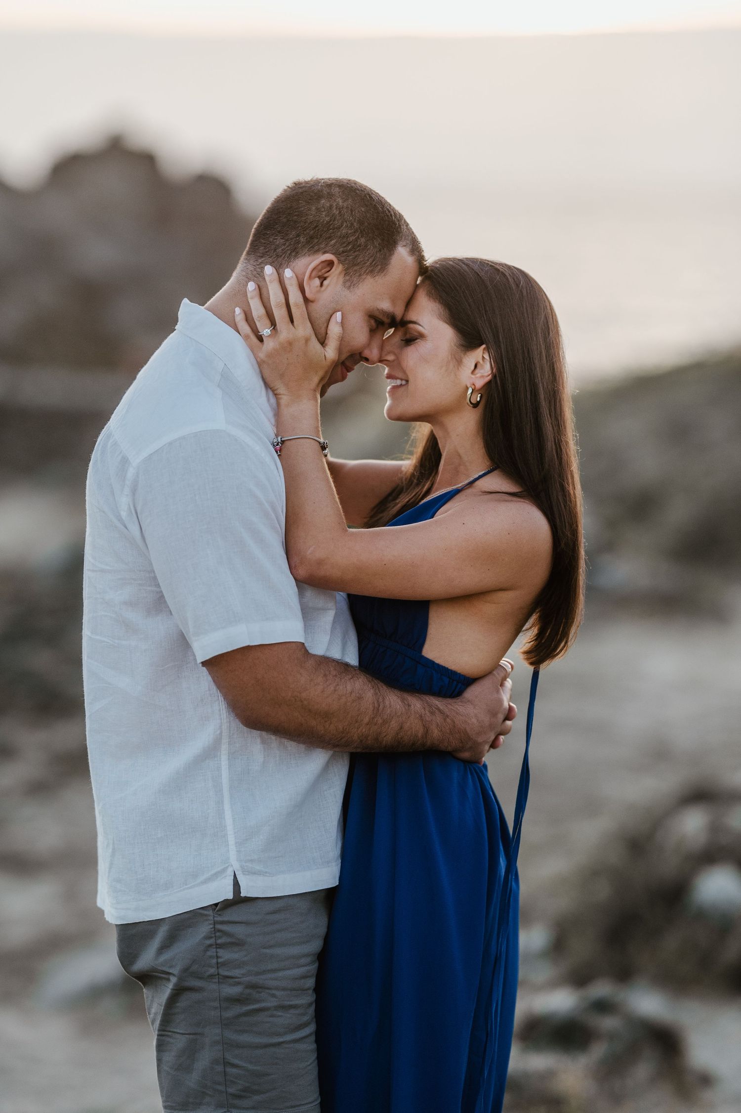 A couple embraces on a rocky beach during sunset, with the woman wearing a navy blue dress and man in white shirt.