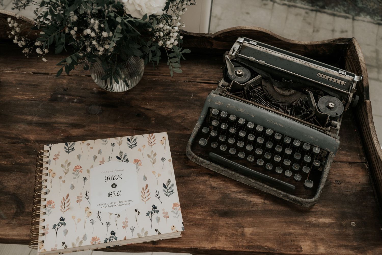 Vintage typewriter and floral notebook on rustic wooden desk with small vase of flowers.