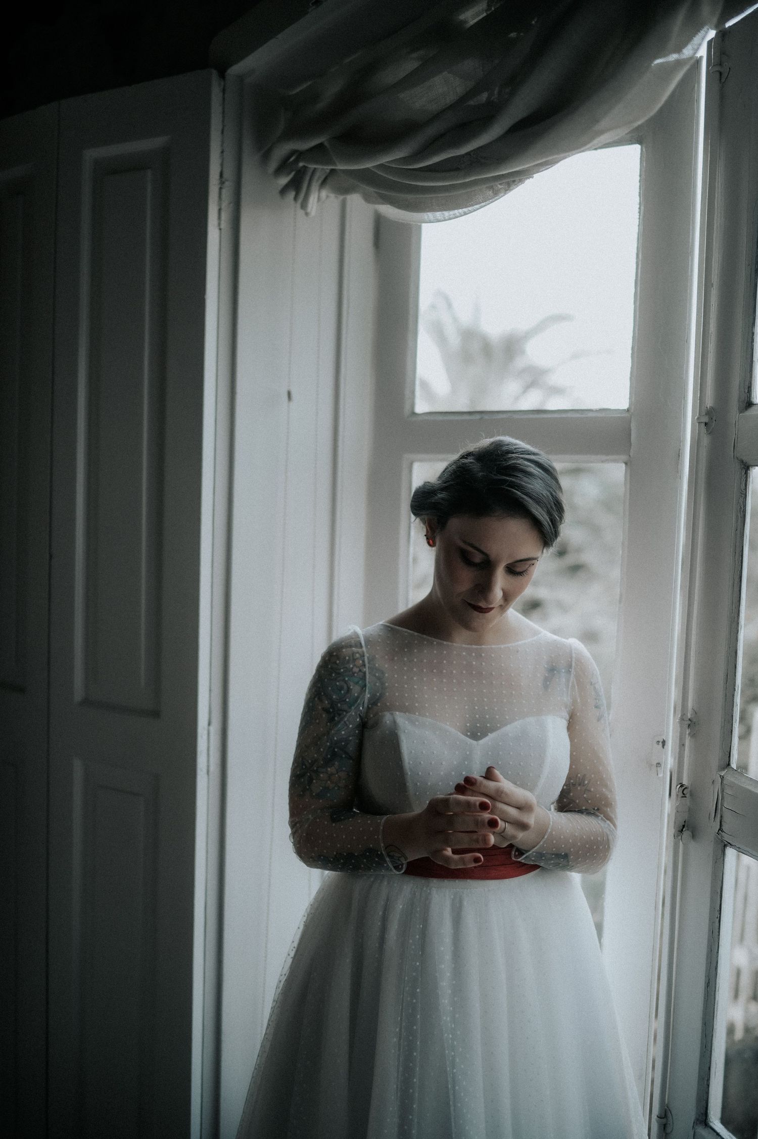 A bride in a white lace wedding dress stands near a window in a moody, atmospheric setting.