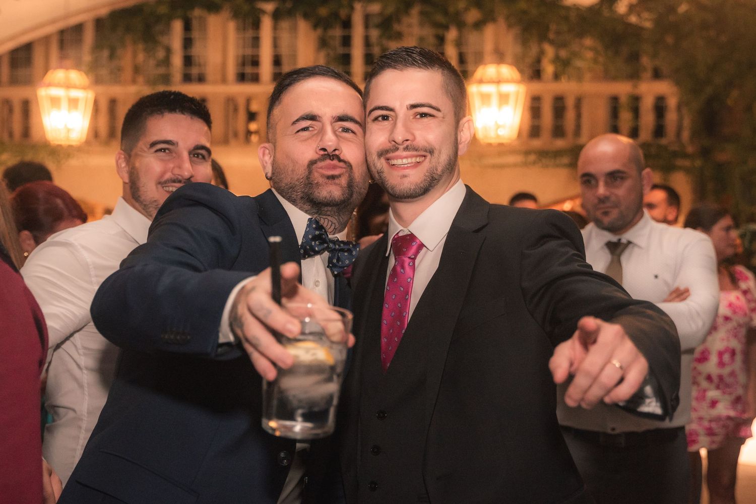 Two men in formal attire smile and point at the camera during an evening celebration at an elegant venue.