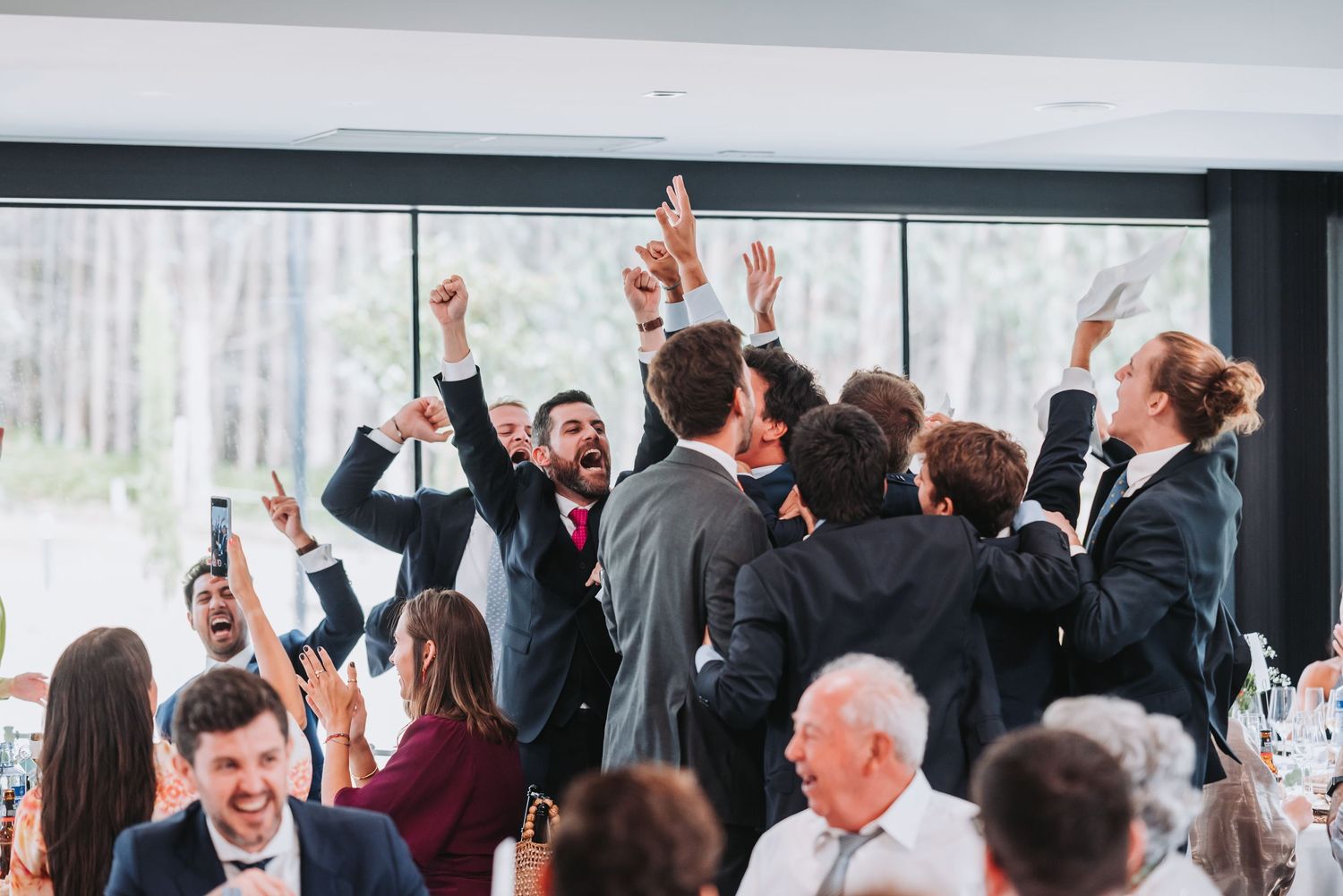 Wedding guests raise their hands and cheer during a celebratory moment at an indoor reception venue.