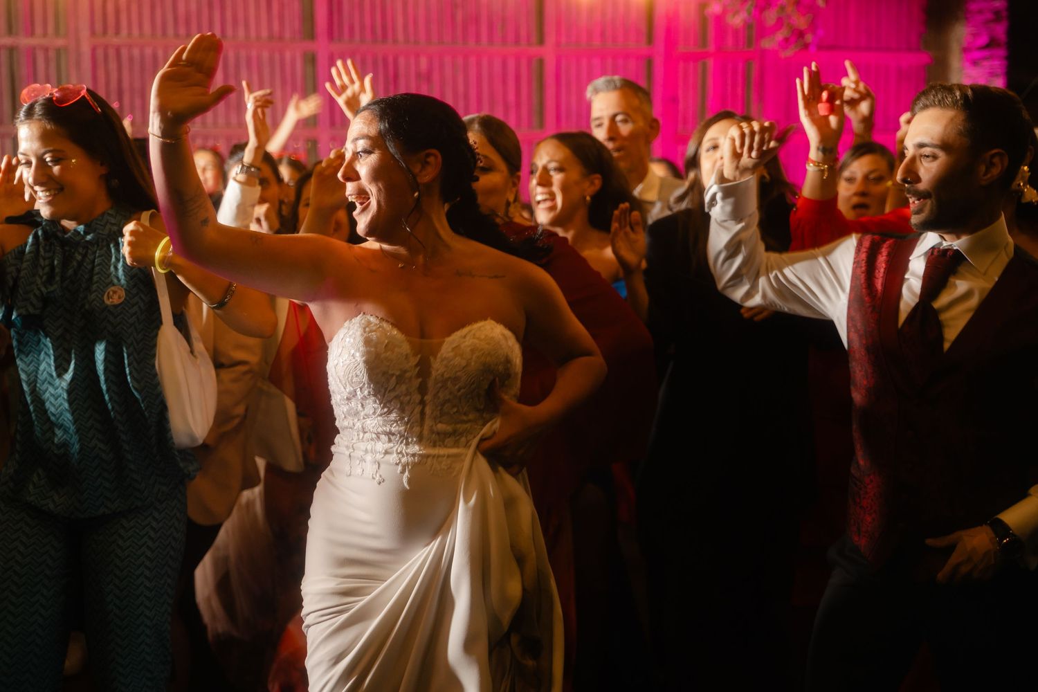 Festive wedding reception crowd dancing under pink lighting and string lights at night.
