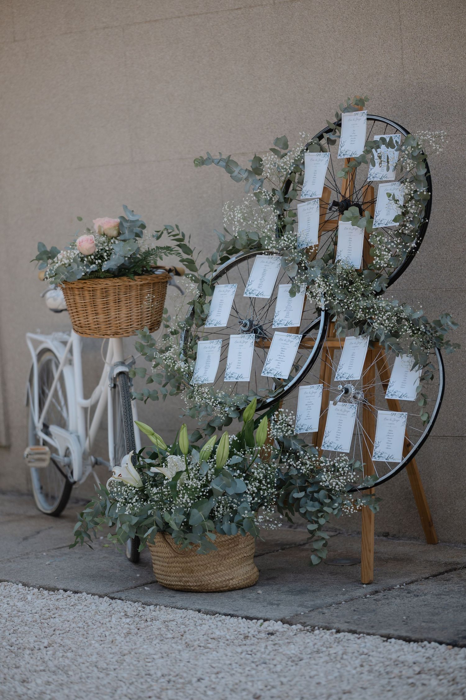 Rustic wedding seating chart display with white cards arranged on wooden ladder alongside wicker baskets and greenery.