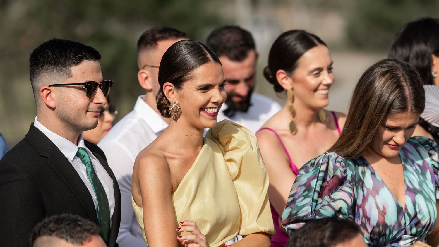 A group of wedding guests smile and laugh together during an outdoor celebration in natural lighting.