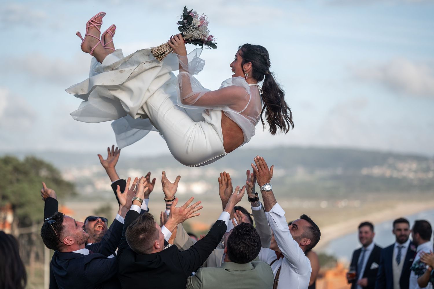 Wedding guests toss a bride and groom into the air during outdoor celebration festivities.