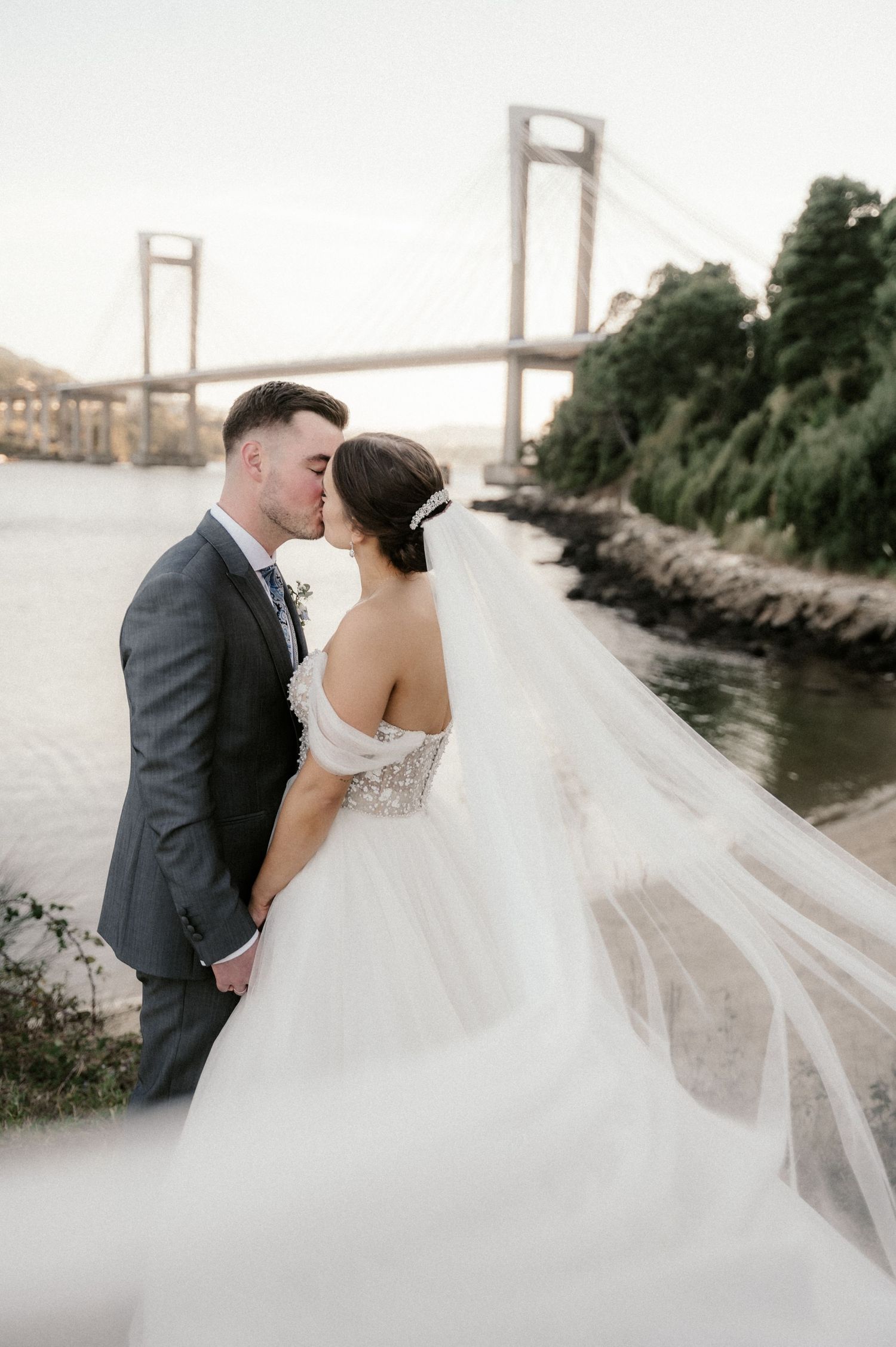 A romantic wedding couple embraces near a bridge with a flowing white veil creating an ethereal scene.