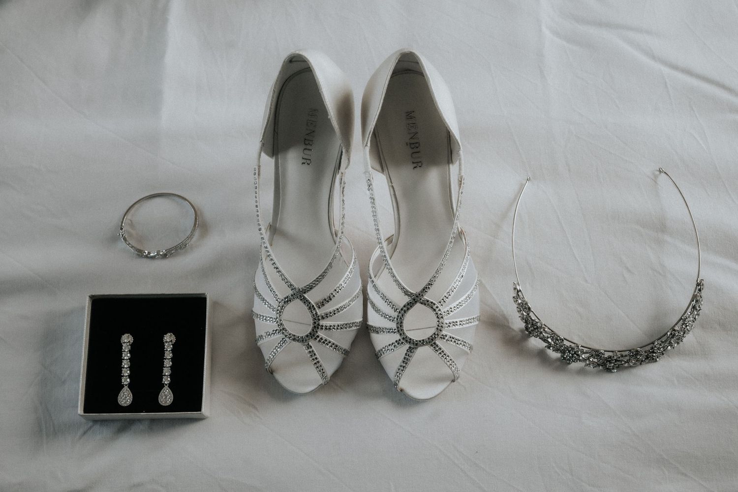 Elegant wedding accessories including white shoes, jewelry, and delicate details arranged on light fabric.