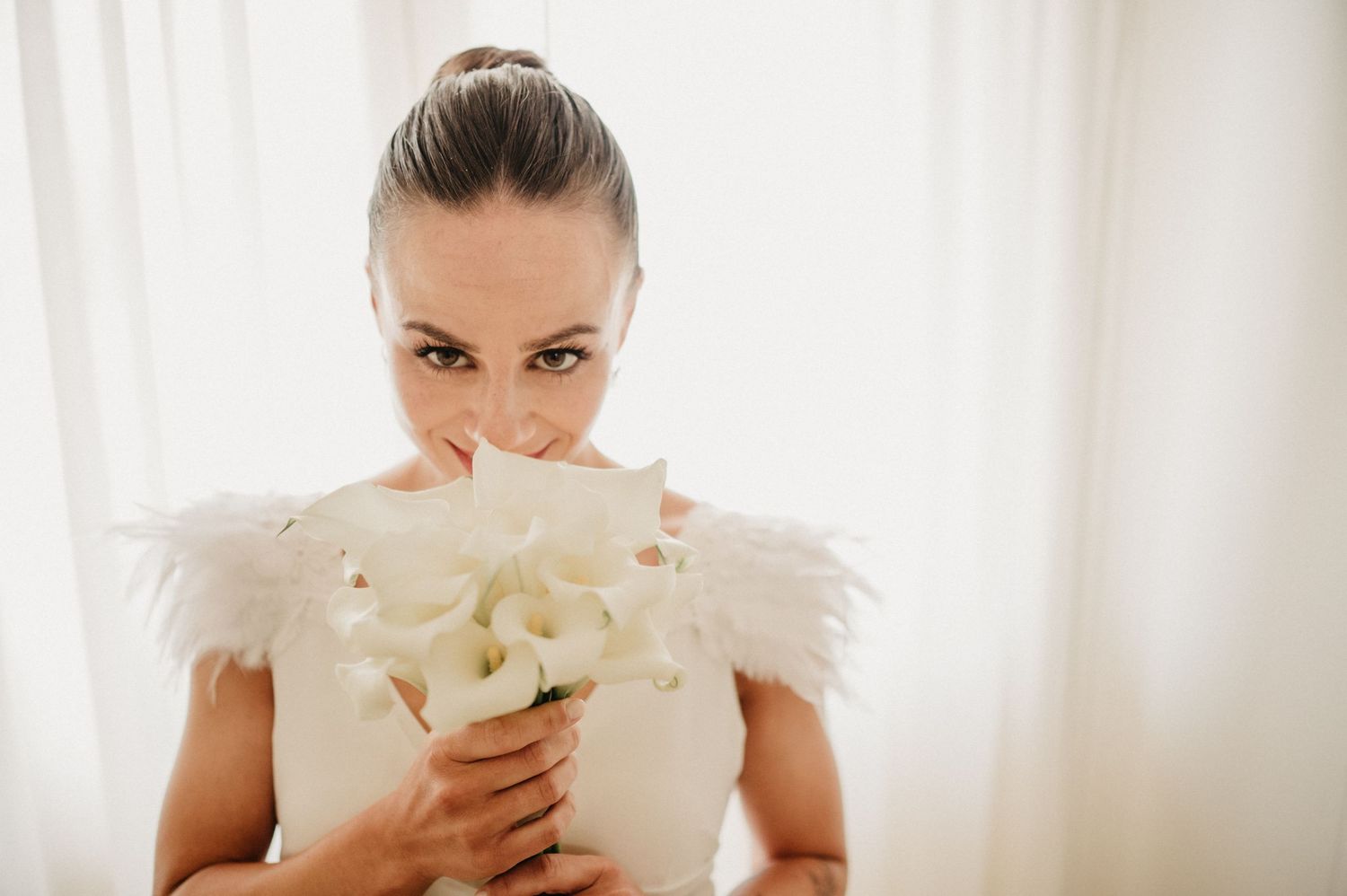 A bride in a white dress with feather details holds a bouquet of white flowers close to her face.