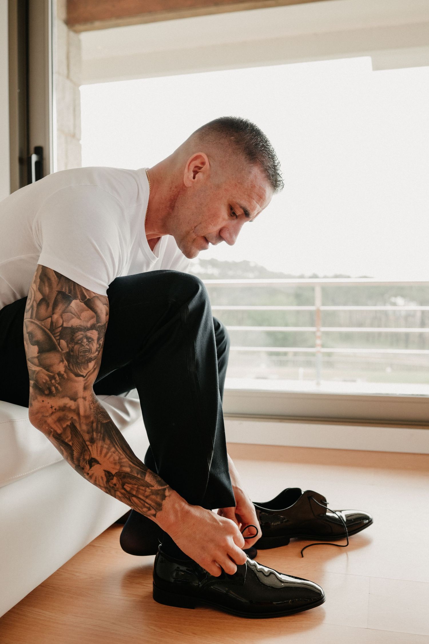 A person with tattooed arms sits on a wooden floor tying black shoes in a bright room by a window.