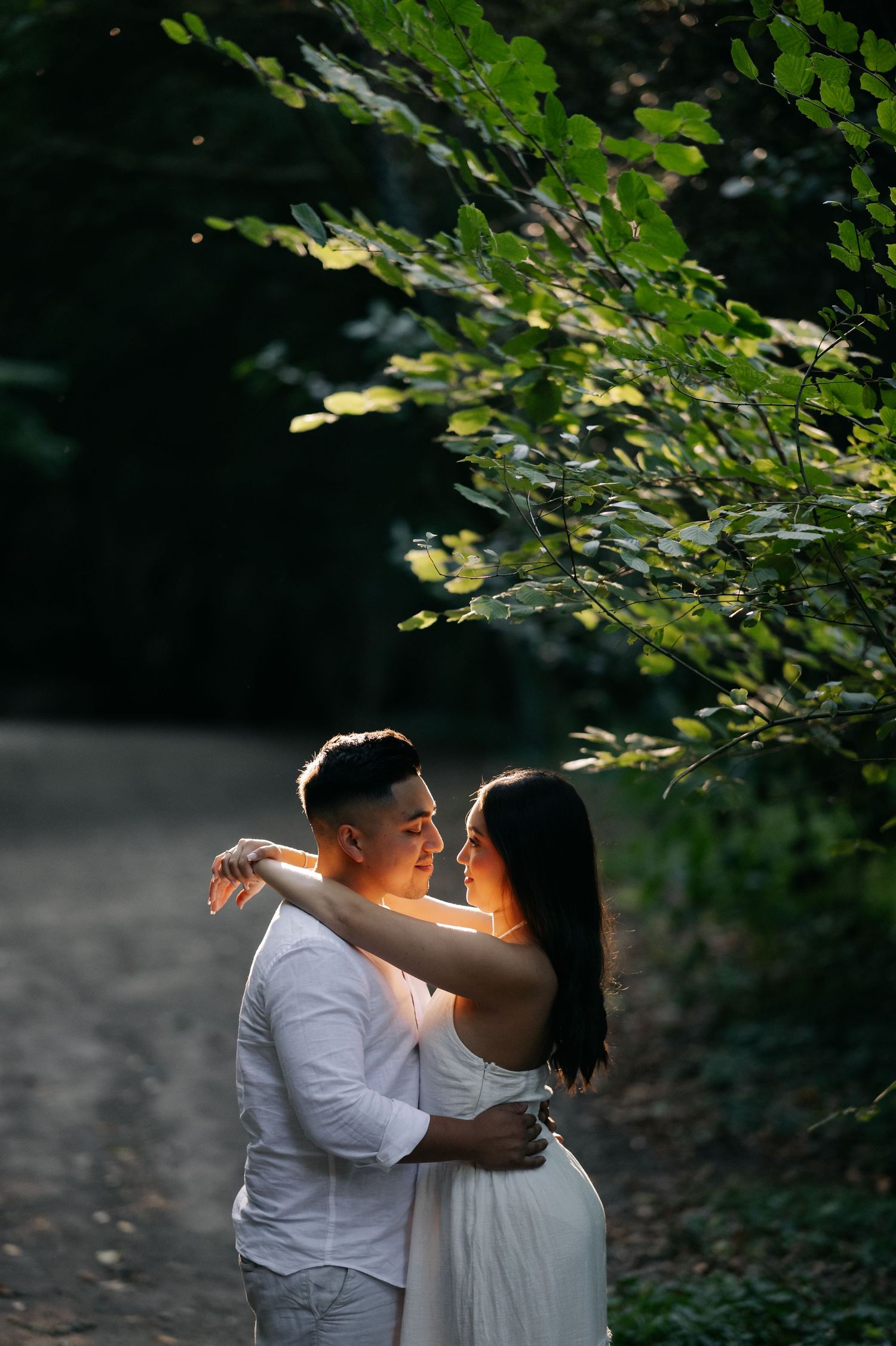 A couple in white attire share a tender embrace under sunlit tree branches on a forest path.