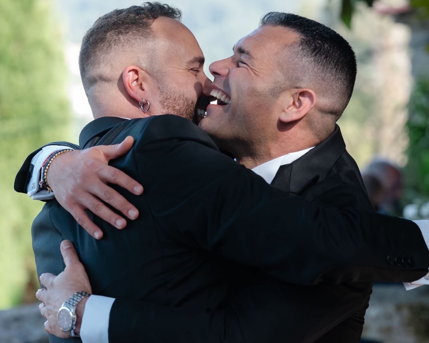 Two people in black suits share a joyful embrace and laugh together during what appears to be a wedding celebration.
