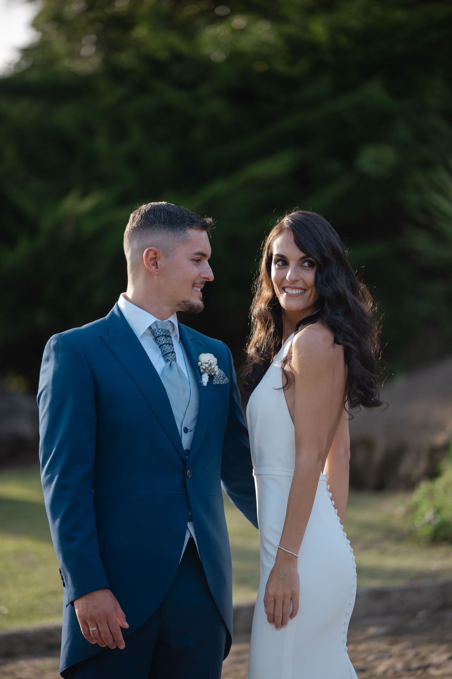 A couple poses together outdoors on their wedding day, with the bride in white and groom in navy blue suit.