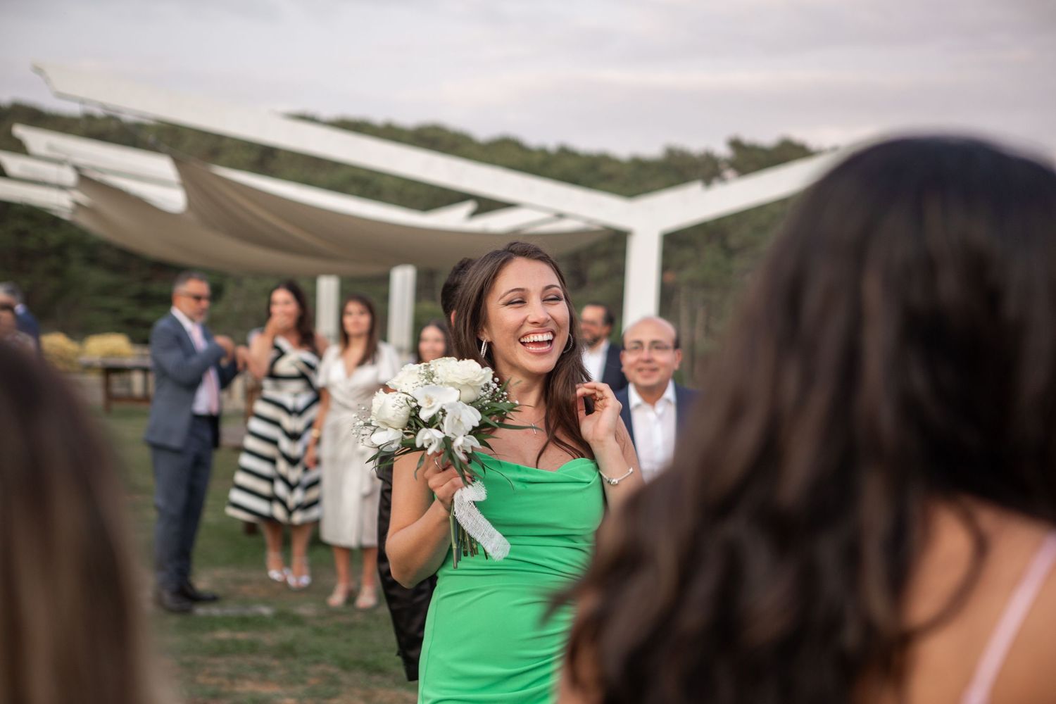 Wedding guest in bright green dress laughs while holding white bouquet at outdoor reception.