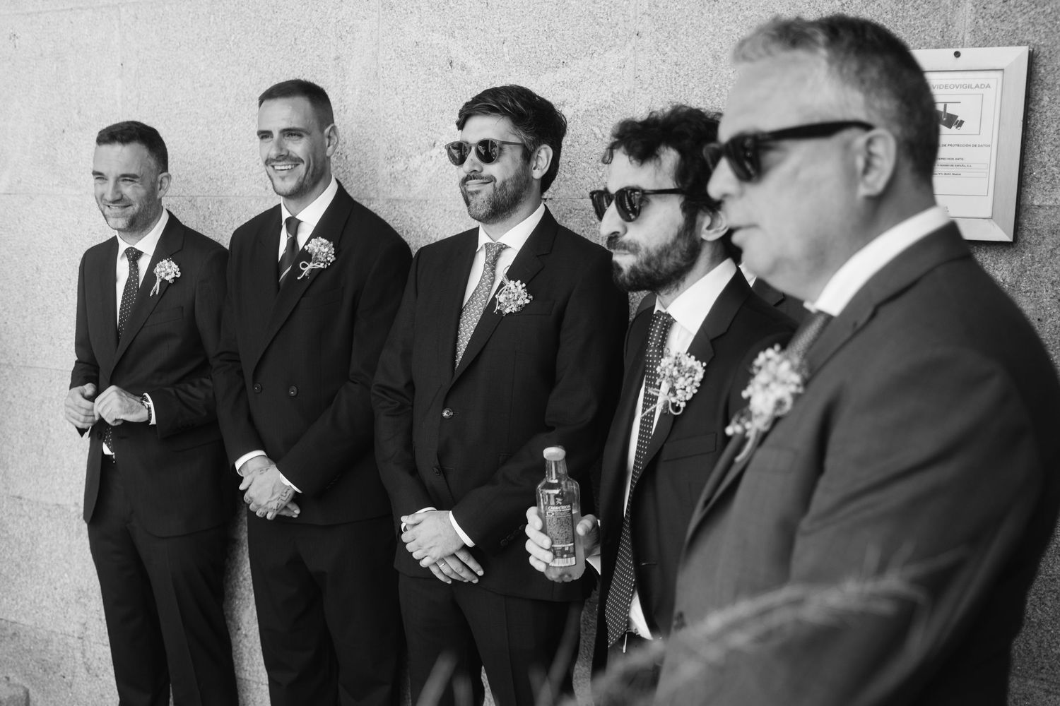 A group of groomsmen in black suits share laughs and drinks during a wedding celebration.