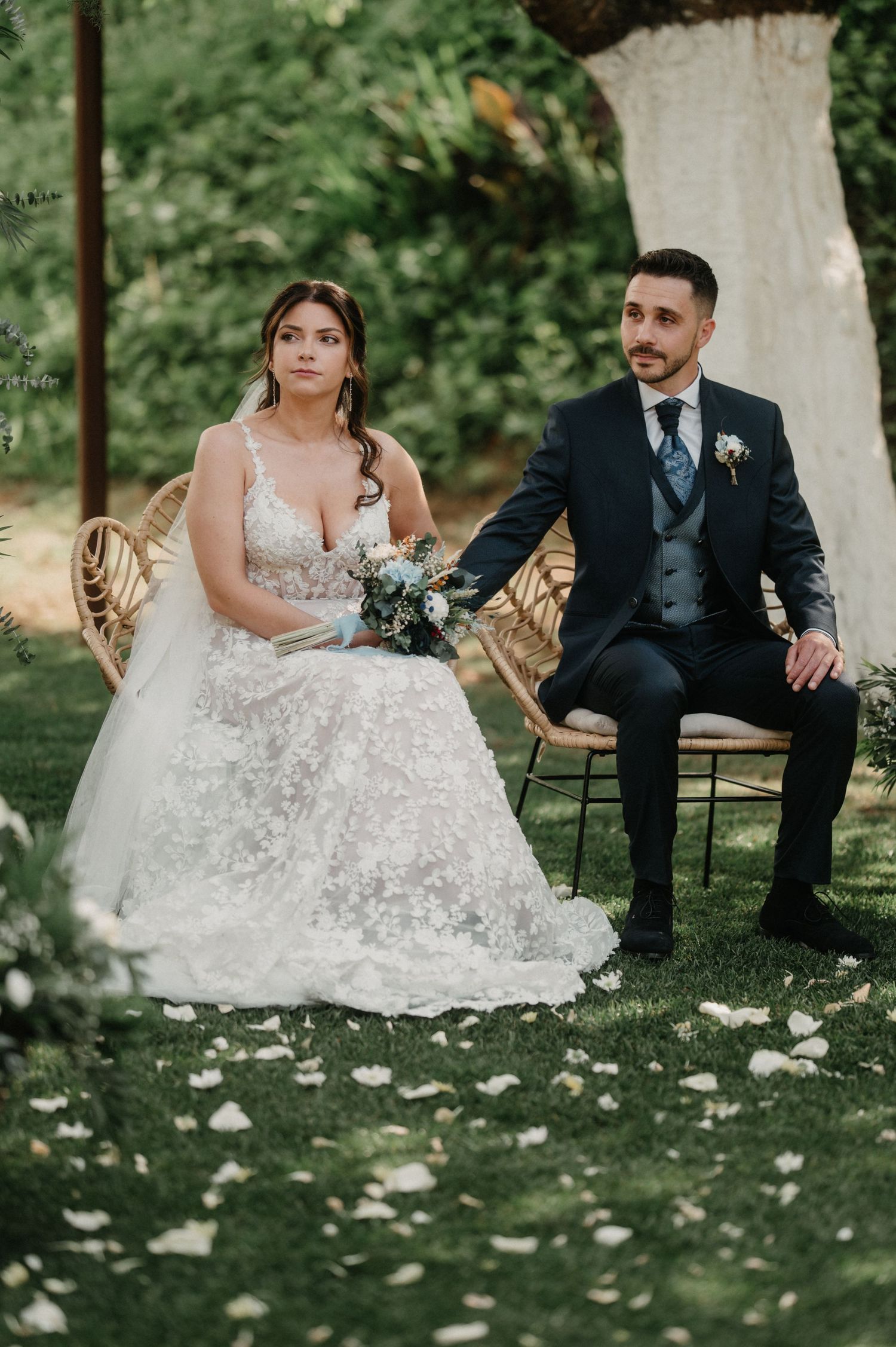 Newlywed couple sits together on vintage chairs in a garden setting with white floral dress details.